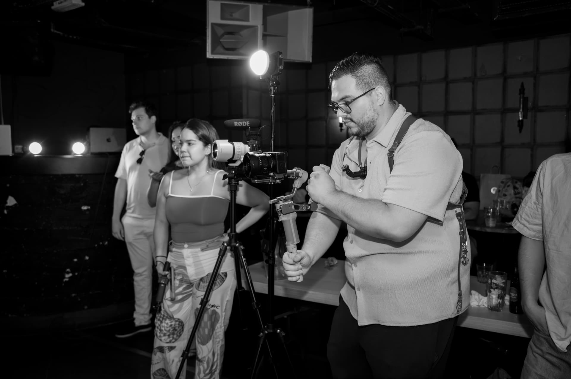 Black-and-white photograph of the studio filming on location: Juan Pablo Arce steadying a camera on a gimbal while María José Ospina stands beside a tripod-mounted camera with a RØDE microphone and a light in a dark venue.