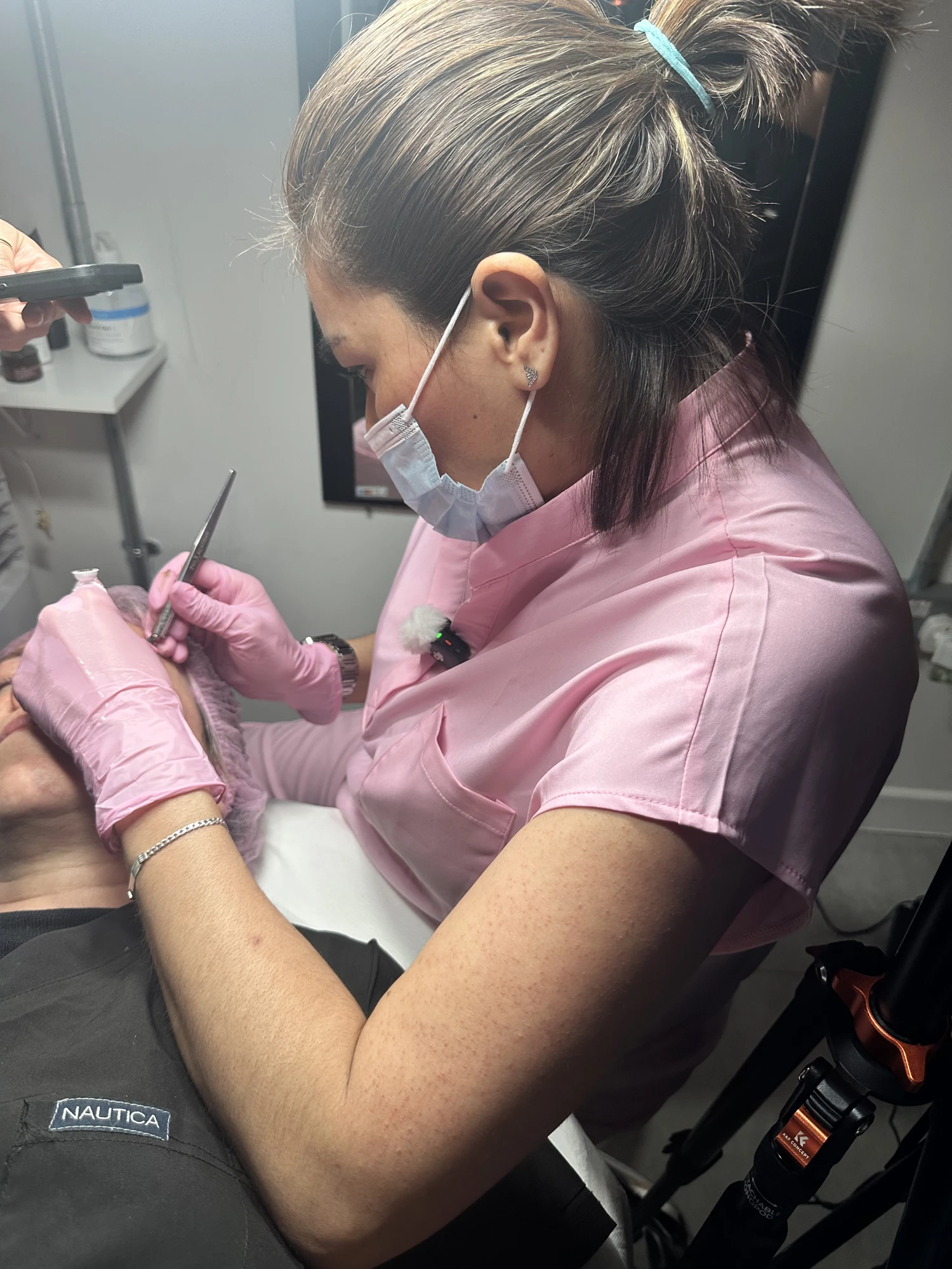 Jenny Agudelo in a pink uniform and pink gloves, working on a client's eyebrows during a treatment, with a microphone clipped on and a camera rig beside her