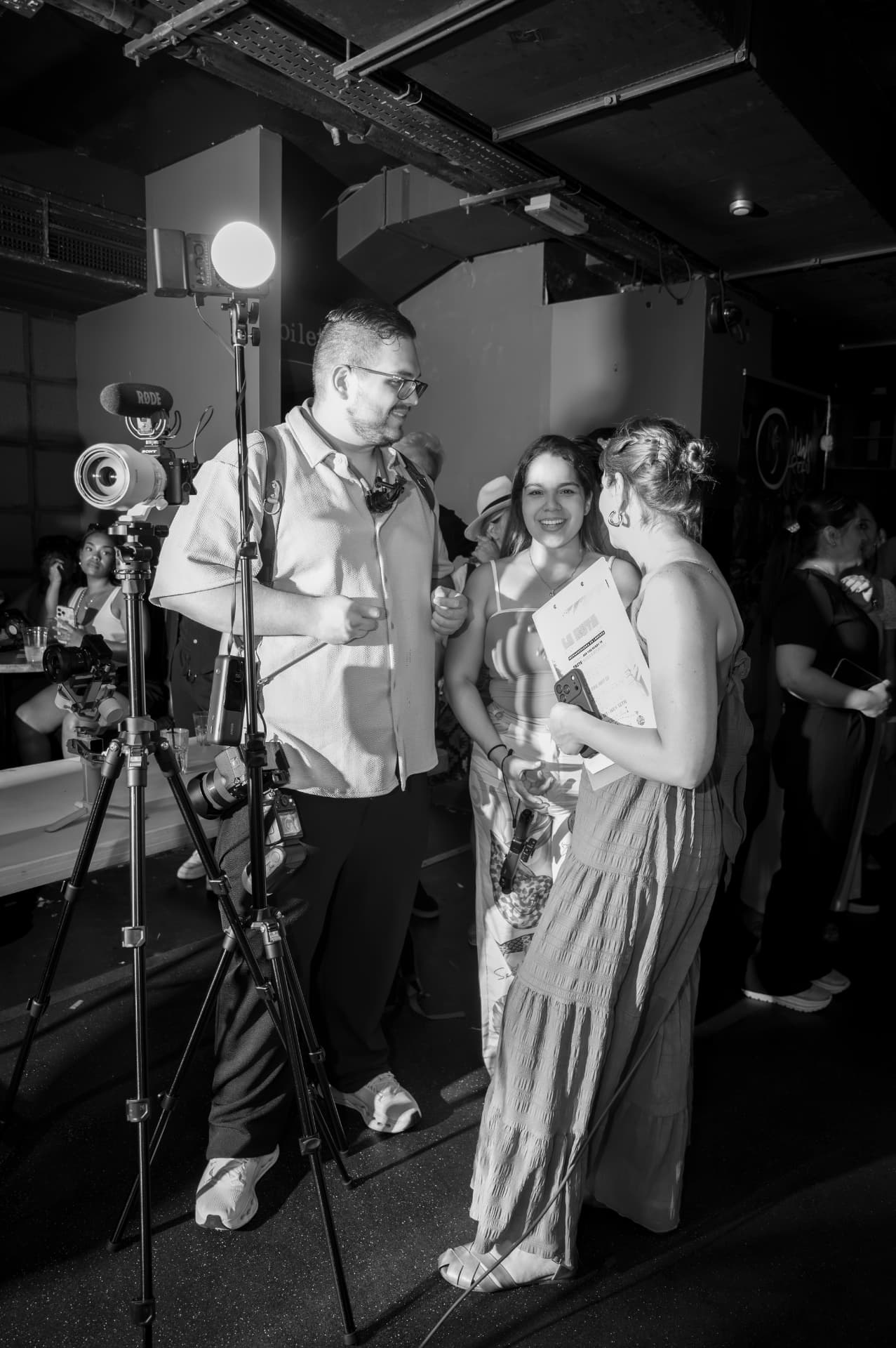 Black-and-white behind-the-scenes photograph of Juan Pablo beside a tripod-mounted camera and production light, speaking with two women at an indoor event.